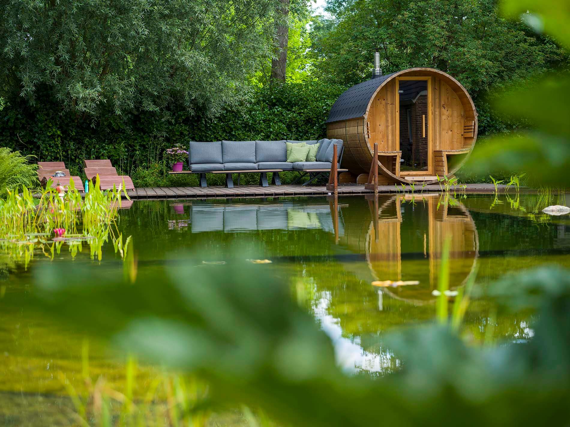 Natuurlijke zwemvijver met sauna, vlonder en loungeplek in een groene tuin.